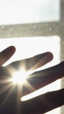 Pencereden süzülen güneş ışığına karşı bir el siluetini yakalayan bir dizi çerçeve. El, güneş ışınlarını engellemek için konumlandırılmış, sanatsal ve düşünceli bir görsel yaratıyor..