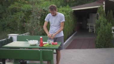 Sakin tavırlı bir adam yemyeşil bir masada, yemyeşil bir bahçede yemek hazırlar. Etrafı yeşilliklerle çevriliyken, sakince sebzeleri doğruyor, huzur ve rahatlama hissi yayıyor.