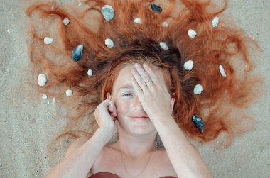 Canlı kızıl saçlı neşeli bir kadın kumlu bir plajda uzanmış, bir gözünü neşeyle eliyle kapatıyor. Küçük taşlarla çevrili, güneşli bir günde kaygısız ve kaprisli bir hava yayar..