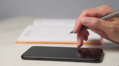 Kalem tutan bir insan, açık bir deftere notlar yazıyor. Yanlarında düz duran siyah bir akıllı telefon var. Üretkenlik, planlama ve teknoloji temaları için mükemmel.