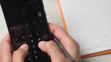 Hesap makinesini elinde kalemle kullanan ellerin yakın plan resmi, arka planda bir defter. Sahne mali hesaplamalar, bütçe hesaplaması ya da iş muhasebe işini içeriyor.