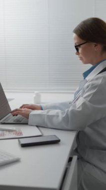 Kendini işine adamış bir sağlık uzmanı modern bir ofis ortamında dizüstü bilgisayar kullanıyor. Beyaz önlük ve gözlük takarak, çalışkan ve profesyonel bir çevreye işaret ederek görevlerine odaklanırlar..