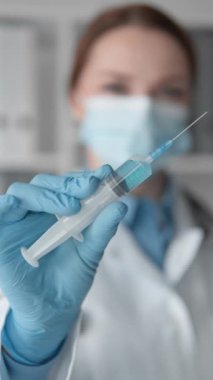 Maske takan ve eldiven takan bir tıp uzmanının elinde aşı dolu bir şırınga var, iğneye hazır. Arkaplanda, klinik ayarların vurgulandığı tıbbi ekipmanlar yer alıyor.
