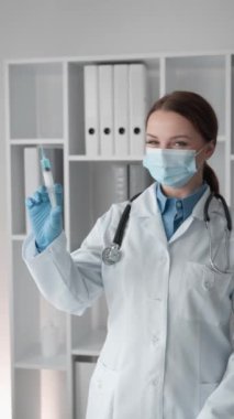 Maske ve eldiven takmış, klinik ortamında şırınga tutan tıp üniformalı bir kadın doktor. Arkaplanda klasörlü raflar ve tıbbi ekipmanlar yer alıyor