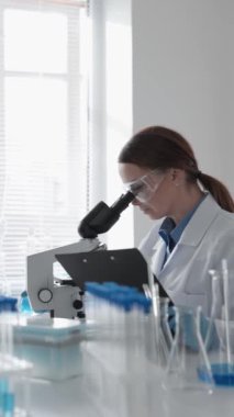 Bir kadın bilim adamı, modern bilimsel araştırma ve yenilikleri gösteren test tüpleri ve bilimsel ekipmanlarla çevrili iyi aydınlatılmış bir laboratuvar ortamında mikroskopla örnekler gözlemliyor..