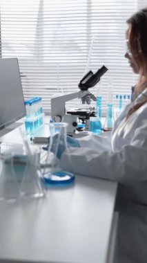 Çağdaş bir laboratuvarda, kadın bir bilim adamı test tüpleri ve çeşitli aletlerle çevriliyken bir mikroskop kullanıyor. Gelişmiş teknoloji onun bitişiğindeki bilgisayar ekranında tasvir ediliyor.