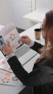 Şirket ofisindeki bir iş kadını mali raporları inceliyor. Veri analizi için dizüstü bilgisayar ve yazılı çizelge kullanıyor. Masası tam bir inceleme için gerekli teknolojiyle donatılmıştır.