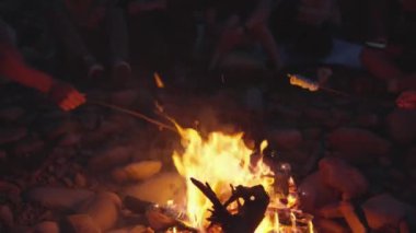 Açık havada bir kamp ateşinin etrafında toplanan arkadaşlar, sıcak ateş gökyüzünü aydınlatırken, yıldızların altında sıcak ve keyifli bir akşam yaratarak atıştırmalıkları kızartıyorlar.