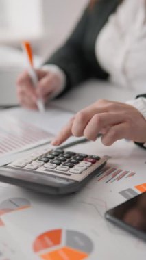 Adanmış bir iş adamı, bir hesap makinesi ve ayrıntılı notlar kullanarak finansal analizler yürütür ve iş stratejisini yönlendiren çok sayıda veri tablosu ve grafik gösterir.