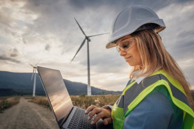 Güvenlik teçhizatı mühendisi rüzgar çiftliğindeki bir dizüstü bilgisayarda çalışarak yenilenebilir enerji ve gelişmiş teknolojinin gezegenimizin sürdürülebilir gelişimindeki temel rolünü sergiliyor.