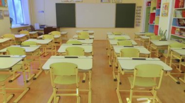 Düzenli masalar, sandalyeler, temiz bir karatahta ve ahşap döşeme içeren parlak, boş bir sınıf. Eğitim temaları, okul ortamı kavramları ve öğrenim alanları için ideal.
