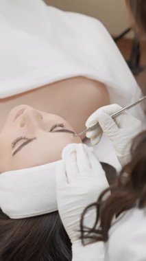 Yüz tedavisi sırasında estetisyen, kafa bandı ve havlu takan rahat bir müşteri için deri temizleme üzerine odaklanır. Bu profesyonel ortamda estetisyen tarafından hassas araçlar kullanılır.