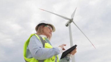 Rüzgar türbinleri olan pitoresk bir manzaranın arka planına karşı, dijital tabletli bir kadın enerji bağımsızlığını sağlamak için rüzgar enerjisinin sürdürülebilir kullanımı için stratejiler geliştiriyor. Yüksek kalite
