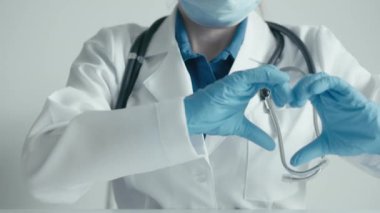 Beyaz önlük ve mavi eldiven giyen bir sağlık uzmanı, şefkati ve şefkati sembolize eden bir kalp hareketi sergiliyor. Steteskop tıbbi bir bağlam gösteriyor..