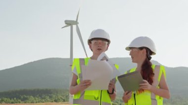 Koruyucu teçhizat ve yüksek görünürlüklü yeleklerde çalışan iki mühendis, yenilenebilir enerji sistemlerinin optimum performans ve güvenliğini sağlamak üzere rüzgar türbini çiftliğindeki bir tablette bulunan belgeleri ve dijital verileri gözden geçirir..