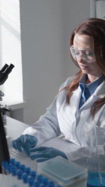 Modern bir laboratuarda, yetenekli bir kadın bilim adamı bir dizüstü bilgisayar ve test tüpleriyle analiz ediyor ve araştırıyor. Güvenlik gözlüğü ve laboratuvar önlüğü takarak deneyler sırasında koruma sağlıyor.