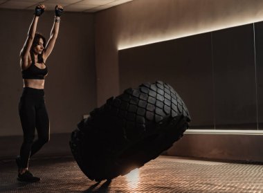 Yoğun bir spor salonunda, odaklanmış bir bayan atlet, güçlü bir lastiği ustalıkla çevirip, sıkı spor yapma diyetinin yoğunluğunu vurgulayarak gücünü ve dayanıklılığını sergiliyor.