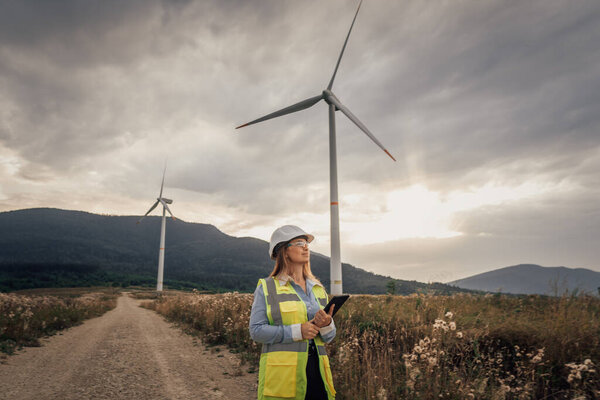 A skilled female engineer in safety gear analyzes data near towering wind turbines in a picturesque landscape, highlighting the essence of renewable energy and sustainable development principles