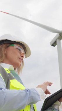 Rüzgar türbinleri olan pitoresk bir manzaranın arka planına karşı, dijital tabletli bir kadın enerji bağımsızlığını sağlamak için rüzgar enerjisinin sürdürülebilir kullanımı için stratejiler geliştiriyor. Yüksek kalite