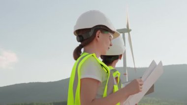 Mühendislerden oluşan bir ekip rüzgâr çiftliğindeki bir rüzgar türbininin belgelerini titizlikle inceliyor. Manzaralı dağların ve açık mavi gökyüzünün ortasındaki sürdürülebilir enerjiye olan bağlılıklarını gösteriyorlar.
