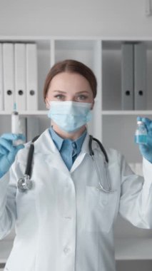 Beyaz önlüklü, maskeli ve eldivenli bir tıp uzmanı aşı şişesi ve şırıngayı modern bir klinik ortamda tutuyor ve aşı ve sağlık hizmetlerinin önemini vurguluyor..