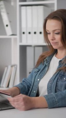 Genç bir kadın modern bir ofiste masa başında otururken tablet kullanıyor. Arkasında raflar ve klasörler var. Sıradan bir iş ortamı. Denim ceket, odaklanmış ifade, profesyonel ayar.