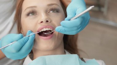 Genç bir kadın bir klinikte diş muayenesinden geçiyor, diş hekimi de oral sağlığını sağlamak için aletler kullanıyor. Yakın çekim prosedürü ve hastaların yüz ifadelerini yakalıyor.