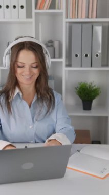 Modern ofiste, mavi gömlekli bir iş kadını işine dalıyor, dizüstü bilgisayar kullanıyor ve kulaklık takıyor. Arka plan kitap rafları ve ofis malzemeleriyle dolu.