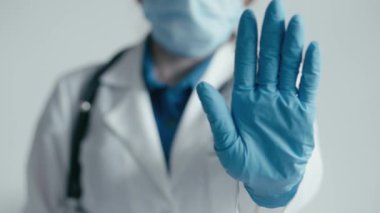 Görüntüde, laboratuvar önlüğü ve steteskop giymiş, maske takmış ve mavi eldiven takmış bir tıp uzmanı, güvenlik önlemleri ve sağlık önlemlerini vurgulayarak, dur işaretinde el kaldırıyor.