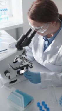 Koruyucu gözlüklü ve laboratuvar önlüklü kadın bilim adamı laboratuvarda mikroskop kullanmaya odaklanıyor. Laboratuvar ekipmanları ve örneklerle çevrili bilimsel araştırma ve analizler yürütüyor..