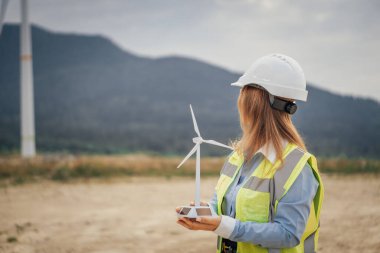 Sert şapka ve güvenlik yeleği giymiş bir kadın mühendis, yeşil enerji ve sürdürülebilir uygulamalarda yeniliği temsil eden yenilenebilir enerji sahasında bir rüzgar türbininin modelini güvenle elinde tutuyor.