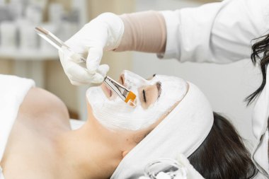 Bir kozmetolog güzellik merkezinde rahatlatıcı bir cilt bakımı sırasında beyaz bir yüz maskesi takmak için bir fırça kullanır. Hasta spa ortamında şımartılıyor, profesyonel güzellik hizmetlerinden zevk alıyor.