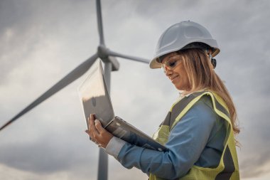 Bir rüzgar türbini sahasında çalışan yetenekli bir kadın mühendis dizüstü bilgisayarını kullanarak yenilenebilir enerji gelişmeleri sergiliyor ve kadınların mühendisliğe hayati katkılarını vurguluyor, gelecek nesillere ilham veriyor.