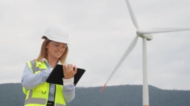 Dijital tabletli bir kadın mühendis, rüzgar türbinlerinin teknik parametrelerini kontrol ederek elektrik santrallerinin sorunsuz çalışmasını ve bunların verimli kullanımını sağlıyor. Yüksek kalite 4k görüntü