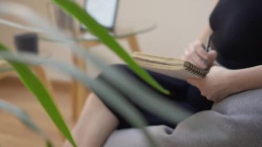 Bir kadın evde rahat bir sandalyeye oturur ve spiral bir not defterine yazmaya odaklanır. Yakınlardaki ahşap bir masada dizüstü bilgisayar görülebiliyor. Bu da ev ofisi ya da yaratıcı çalışma alanı ayarlarını gösteriyor..