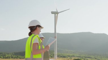 Yüksek görünürlük yelekleri ve sert şapkalar giyen bir grup mühendis, yeşil enerji teknolojisiyle sürdürülebilirlik üzerine çalışan bir rüzgar türbini projesi için açık havada birlikte çalışıyorlar.