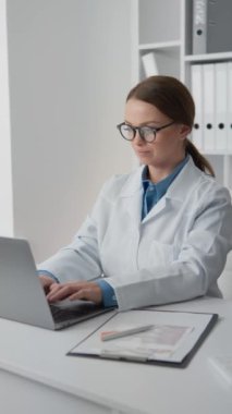 Çağdaş bir tıp kliniğinde, beyaz laboratuvar önlüğü ve gözlüklü bir kadın doktor özenle bir dizüstü bilgisayar üzerinde çalışıyor. Masa düzenli, tıbbi kayıtlar ve evraklar düzenli.