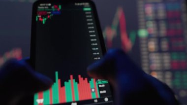 Gerçek zamanlı borsa verilerini gösteren bir akıllı telefonun yakın çekimleri. Eller ekranla etkileşime girer, dalgalanmaları ve eğilimleri analiz eder. Arkaplanda ek ticaret grafikleri gösteriliyor.