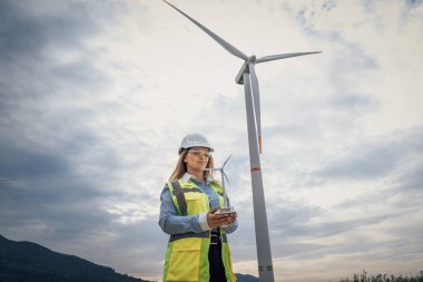 Güvenlik teçhizatında yetenekli bir kadın mühendis, yenilenebilir enerji ve kadınların mühendislik ve sürdürülebilir uygulamalardaki yetkilerini vurgulayan güçlü bir rüzgar türbininin yanında gururla duruyor.