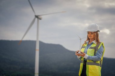 Sert şapka ve güvenlik yeleği giymiş bir kadın mühendis, büyük bir rüzgar çiftliğinin yanında dururken minyatür bir rüzgar türbini modelini inceliyor ve yenilenebilir enerji gösteriyor.
