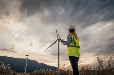 Parlak güvenlik yeleği ve sert şapkalı yetenekli bir kadın teknisyen, günbatımında uzun bir rüzgar türbini inceleyerek, barışçıl bir kırsal çevre için yenilenebilir enerjinin önemini vurguluyor.
