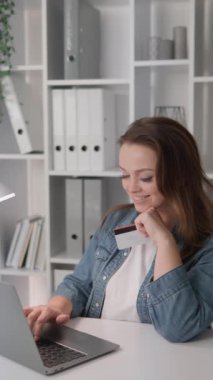 Modern bir çalışma ortamında, bir ev ofisinde genç bir kadının dizüstü bilgisayar ve kredi kartı kullandığı görülüyor. Klasörlü raflar ve dekoratif öğeler çağdaş bir ayar oluşturur