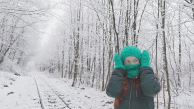 Şık yeşil şapkalı ve eşarplı neşeli bir kadın, karlı bir arazide poz veriyor, kış tatilinin ve bayram kutlamalarının özünü yakalıyor, neşe ve heyecan uyandırıyor.