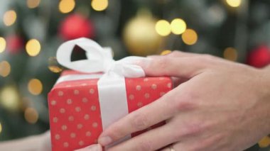 Beyaz kurdeleli canlı kırmızı bir hediye kutusu renkli süsler ve sıcak ışıklarla süslenmiş neşeli bir Noel ağacının önünde gururla sunuluyor ve neşeli bir atmosfer yaratıyor.