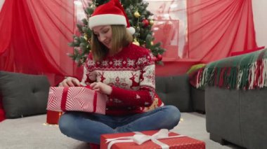 Neşeli bir tatil sahnesinde, Noel Baba şapkası ve şen süveter giyen genç bir kadın, güzel dekore edilmiş bir ağacın altında neşe içinde Noel hediyelerini açıyor ve mevsimlerin büyüsünü arttırıyor.