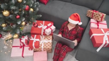 Neşeli bir Noel sahnesinde renkli pijama ve Noel Baba şapkası giymiş neşeli bir çocuk şaşaalı bir şekilde süslerle ve süslemelerle dolu parlak bir ağacın altında hediyeler arasında oynar.