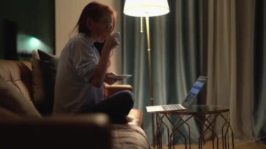 Rahat bir kadın, evinde rahat bir şekilde rahat mobilyalarla dolu huzurlu bir oturma odasında laptopuyla çalışırken lezzetli bir fincan kahvenin tadını çıkartıyor.