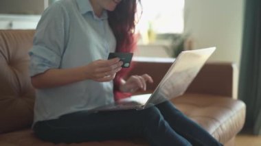 Genç bir kadın, elinde kredi kartıyla internetten alışveriş yapmaya odaklanmış şık kanepesinde oturuyor. Konforlu, modern yaşam alanı onun keyifli ve uygun deneyimini arttırıyor.