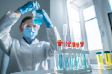 Beyaz önlüklü genç bir bilim adamı renkli test tüpleriyle karmaşık deneyler yapıyor. Kapsamlı bilimsel araştırmalar için hassasiyet, adanmışlık ve özen gösteriliyor.