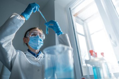 Laboratuvar önlüğü ve koruyucu giysi giyen kendini adamış bir bilim adamı, keskin ve güvenli bir laboratuvarda pipetler ve cam eşyalar ile hassas bir deney gerçekleştiriyor.